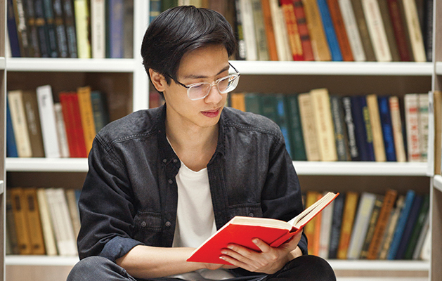 Bookworm Concept. Creative asian male student sitting on the floor and reading a book at library, free space