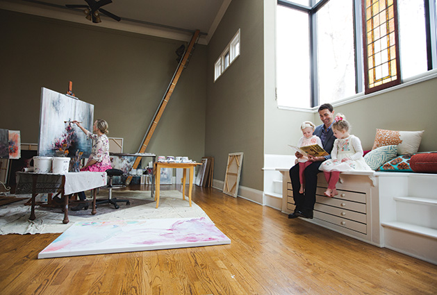 Artist Jillian Lee painting while her husband reads a book to their children.