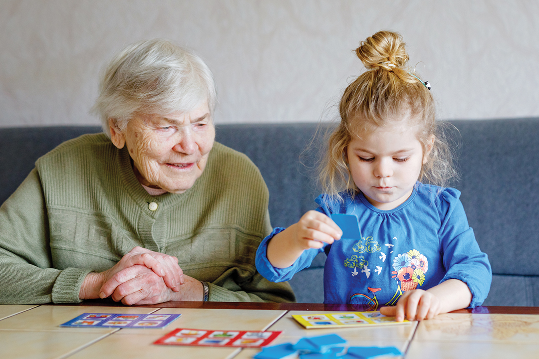 Beautiful toddler girl and grand grandmother playing together pictures lotto table cards game at home. Cute child and senior woman having fun together. Happy family indoors.