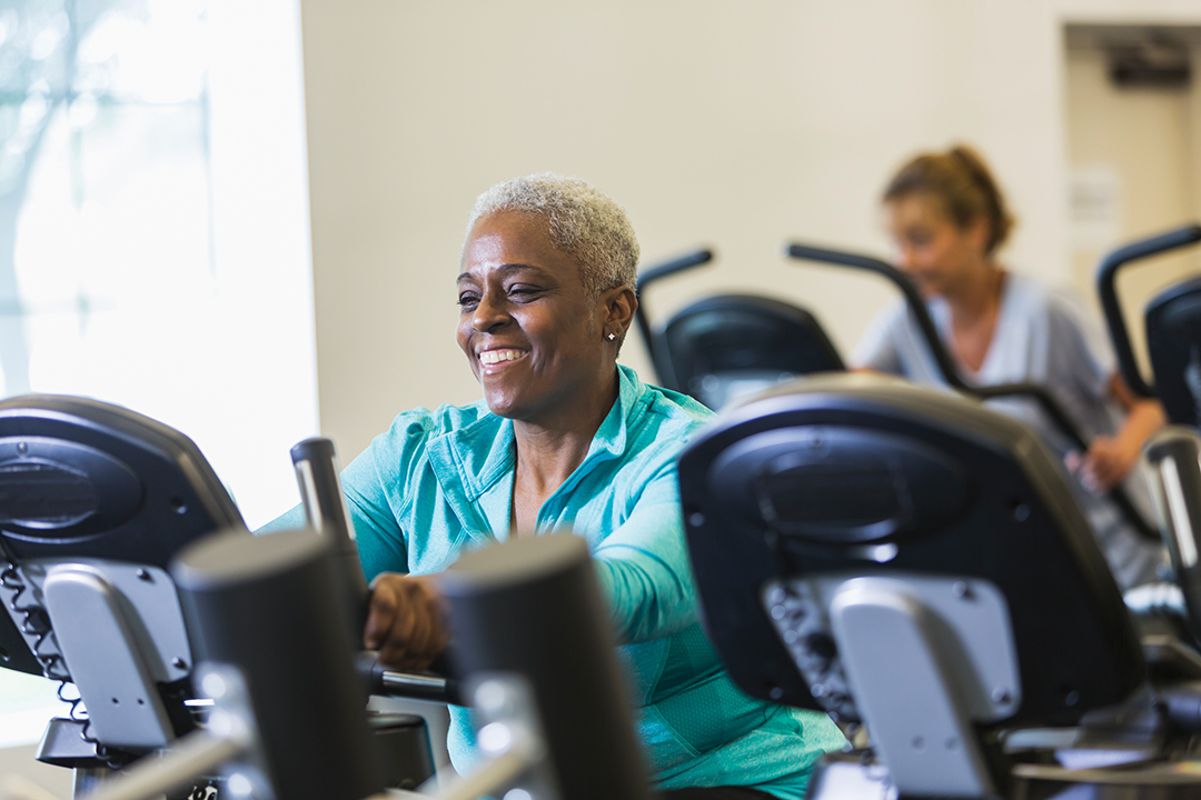 Happy senior African American woman (60s) at health club riding exercise bike, smiling.