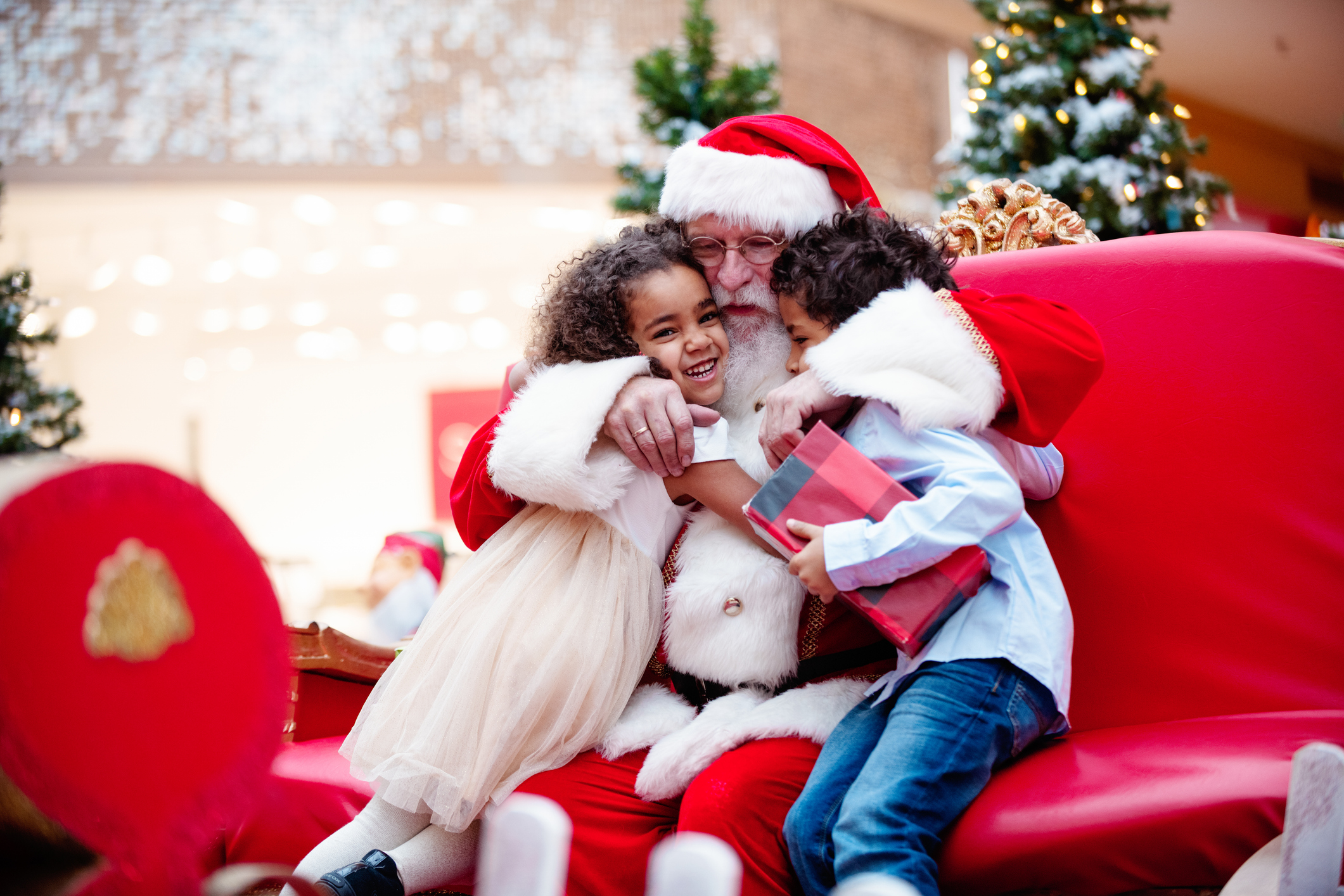 Multi-Ethnic family shops at Shopping Mall during Christmas Time with Santa Claus. Santa Claus is asking the children what they want for Christmas. Brother and sister happy to see Santa Claus. They are hugging. Santa has arms around children. Boy has gift in his hands. Photo was taken in Quebec Canada.