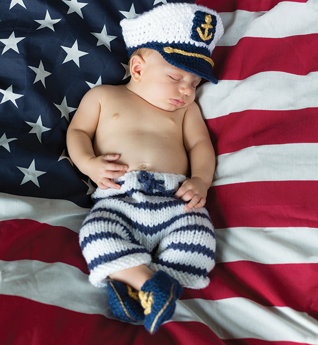 Baby in knit sailor's hat and pants with an American flag in the background.