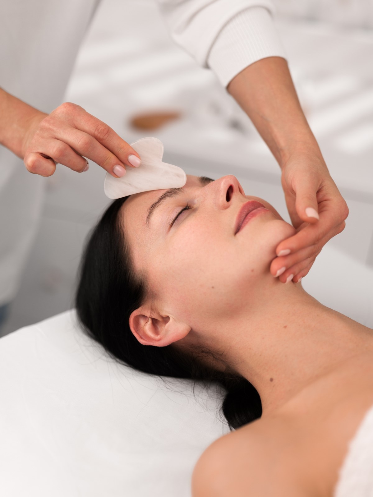 From above of crop unrecognizable cosmetician using gua sha tool while massaging face of peaceful young female client lying on couch with closed eyes