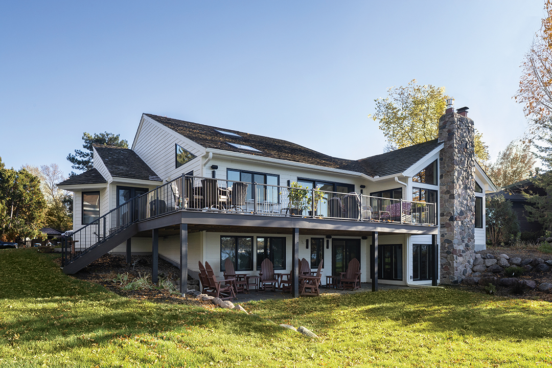 The exterior represents a dramatic shift from dark midcentury stains to a bright, high-contrast facade that feels expansive against the natural shoreline. This fresh aesthetic is anchored by preserved stonework on the towering chimney, maintaining a physical link to the property’s history while updated windows and grand sliding doors frame the lake as the central feature. Outdoor living is elevated by a sprawling wraparound deck, featuring vertical cable railings for uninterrupted views and a clever pass-through window with a bar-style countertop, explains Kimberly Fletcher, lead designer at Boyer Building Corporation. This design erases the boundary between the kitchen and the outdoors, supporting a seamless flow of activity for long days spent by the water.