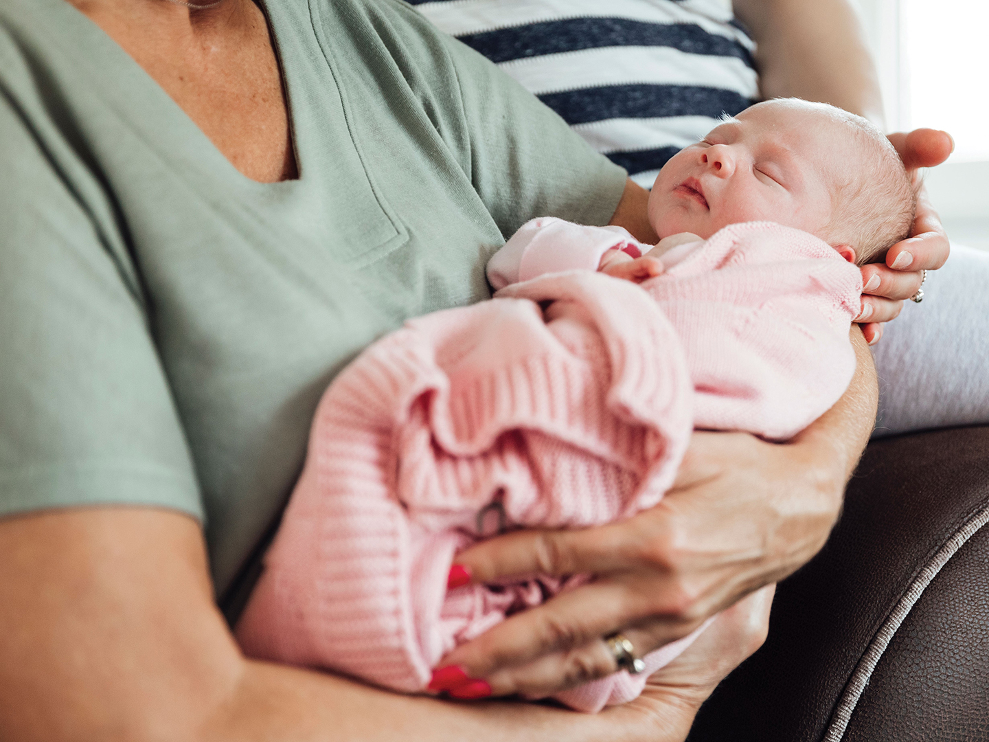 A close-up of a grandmother holding her newborn granddaughter on her lap, with her daughter beside her, the grandmother is wearing a facemask.